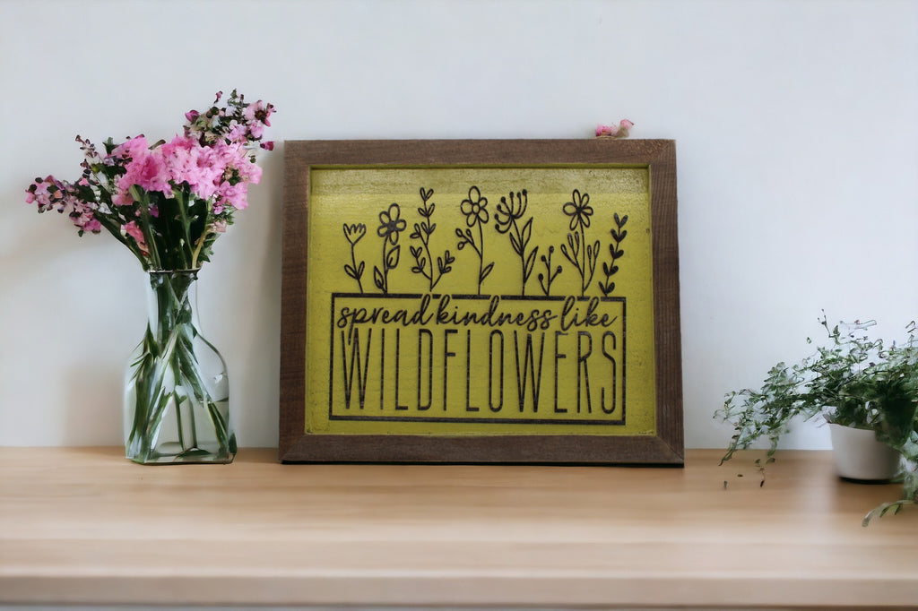 A yellow sign with beautiful text that reads 'Spread Kindness Like Wildflowers,' surrounded by illustrations of delicate wildflowers."